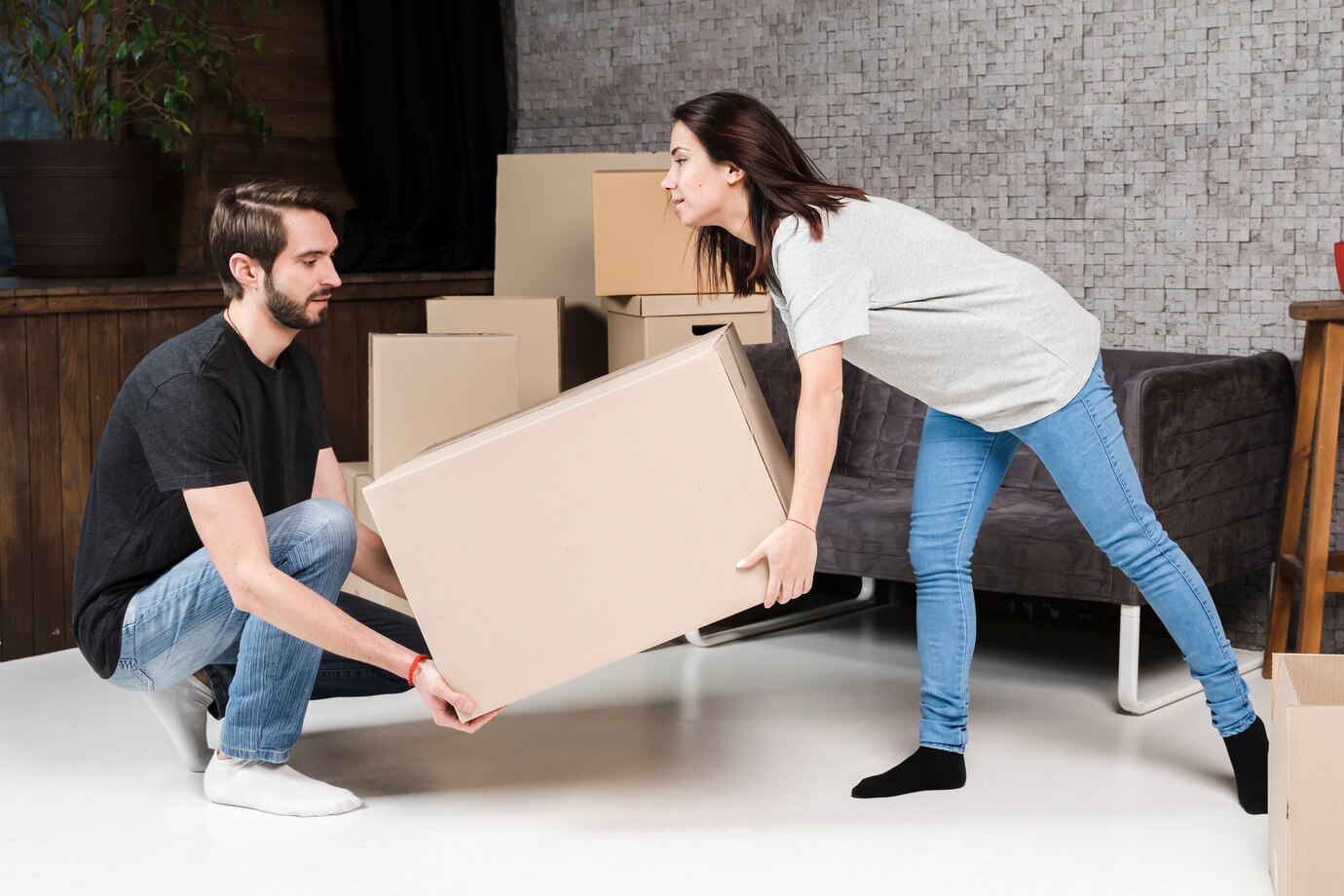 Two People Lifting A Cardboard Box In A Room With Stacked Boxes And Furniture