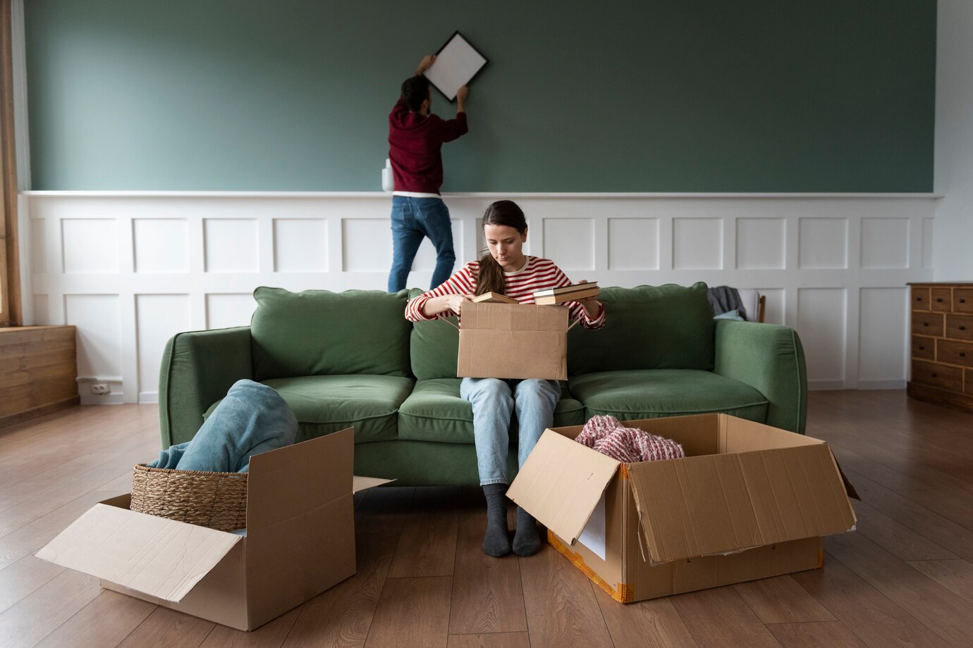 Two People Unpacking Boxes In A Room With A Green Sofa And Teal Wall