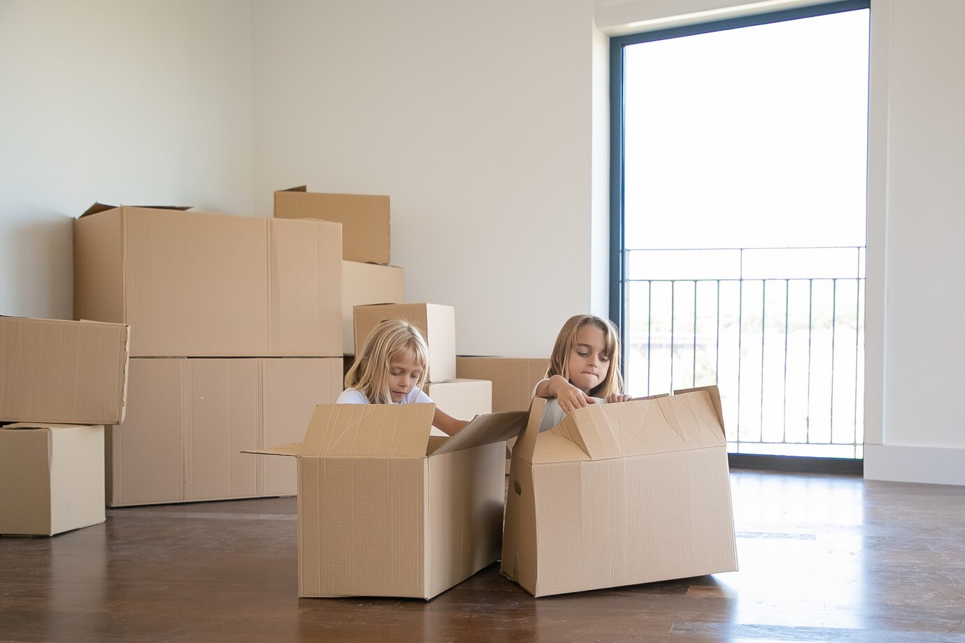 Two Children Playing In Cardboard Boxes In A Room With Stacked Boxes And A Window